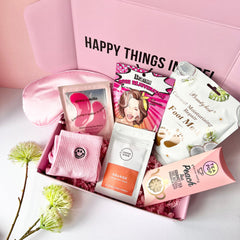 A pink Jumbo Pamper Box open to display its contents, which include a pair of pink smiley socks, a coffee scrub pouch, a satin sleep mask, a Korean face mask sheet, a coconut foot pack, a herbal tea pop and pink eye patches. All arranged neatly inside the box.