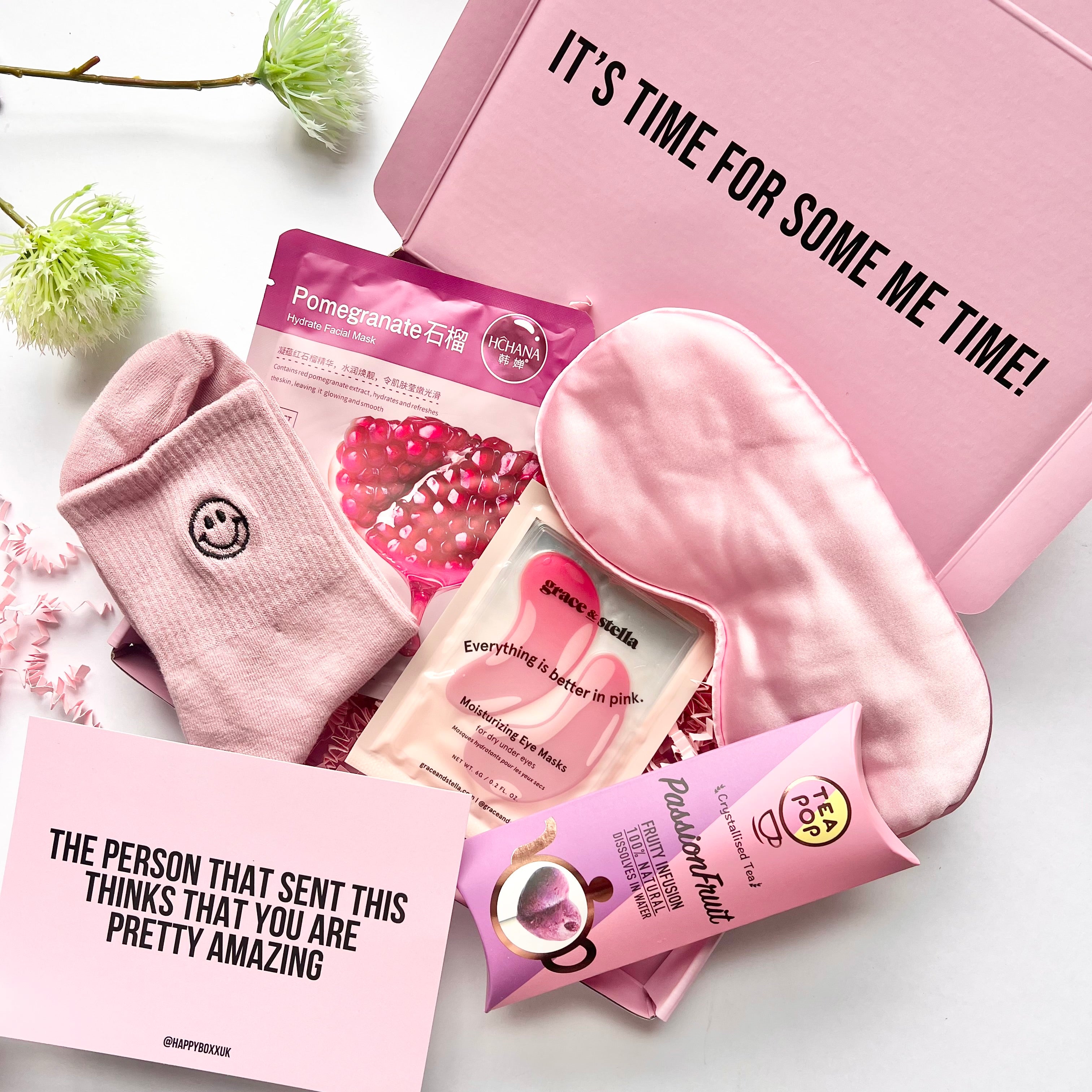 Pink self-care products including satin sleep eye mask, Korean face mask, Grace & Stella moisturising eye patches, herbal tea pop and pink smiley face socks with a greeting card reading 'the person that sent this thinks that you are pretty amazing'. All placed in a pink gift box.