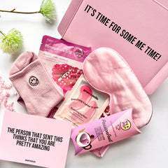 Pink self-care products including satin sleep eye mask, Korean face mask, Grace & Stella moisturising eye patches, herbal tea pop and pink smiley face socks with a greeting card reading 'the person that sent this thinks that you are pretty amazing'. All placed in a pink gift box.