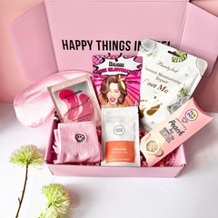 A pink Jumbo Pamper Box open to display its contents, which include a pair of pink smiley socks, a coffee scrub pouch, a satin sleep mask, a Korean face mask sheet, a coconut foot pack, a herbal tea pop and pink eye patches. All arranged neatly inside the box.