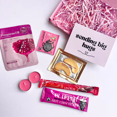 Flat lay image of assorted items including a greeting card reading 'sending big hugs', pink tealights, chocolate bar and hot chocolate sachet on a white background.
