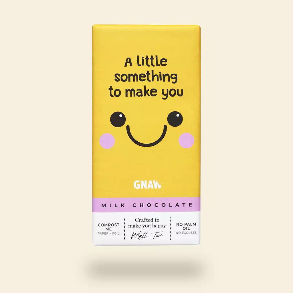 A yellow chocolate bar packaging with a smiling face and the text 'A little something to make you' on top. The brand 'GNAW' is visible at the bottom with the product description 'Milk Chocolate' and additional text indicating compostable packaging.