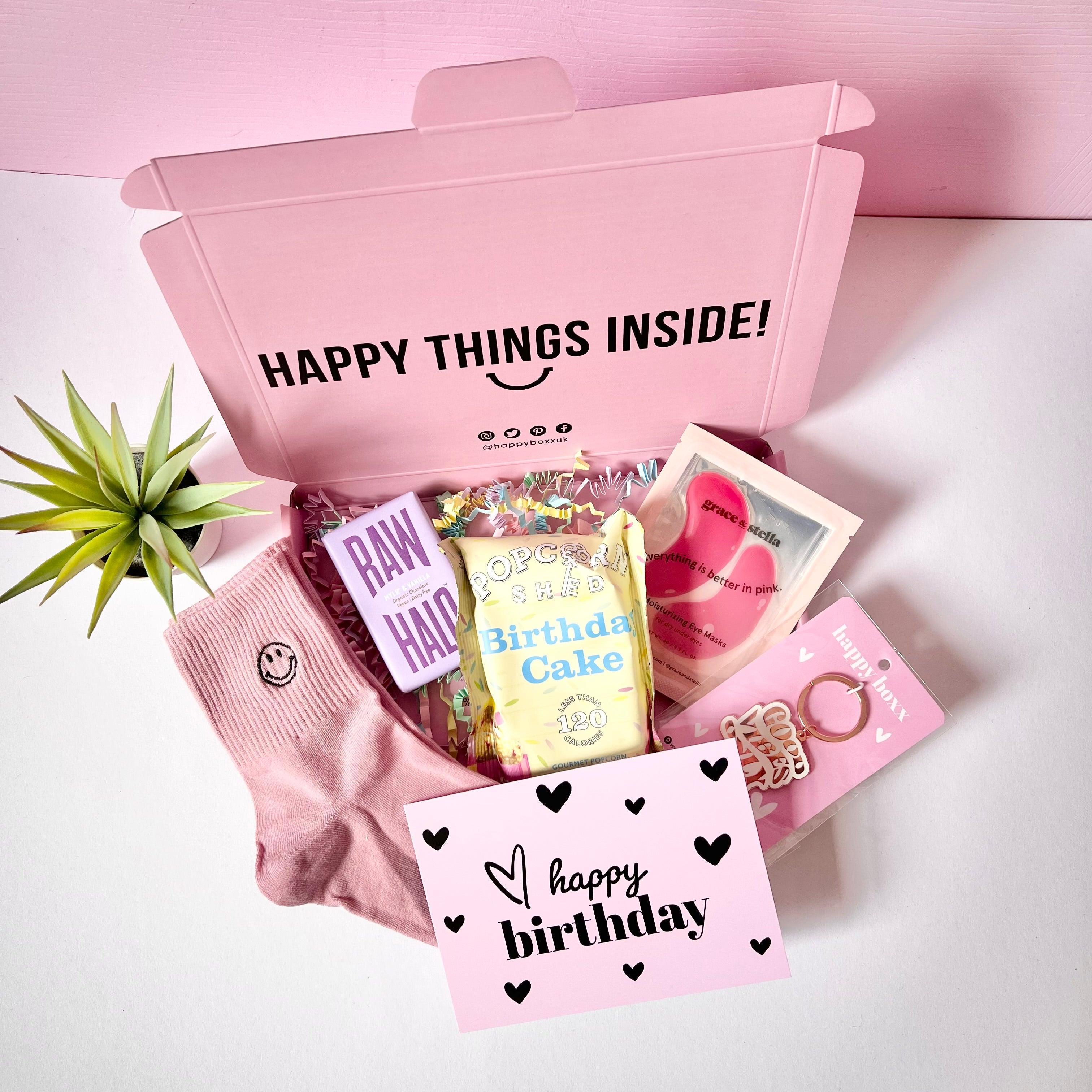 A pink gift box open to reveal various birthday gifts including pink smiley face socks, a 'happy birthday' greeting card, eye patches, keyring, popcorn, and chocolate bar. The box is labeled 'HAPPY THINGS INSIDE!' and there's a 'happy birthday' card visible inside the box.