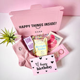 A pink gift box open to reveal various birthday gifts including pink smiley face socks, a 'happy birthday' greeting card, eye patches, keyring, popcorn, and chocolate bar. The box is labeled 'HAPPY THINGS INSIDE!' and there's a 'happy birthday' card visible inside the box.