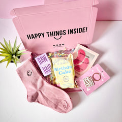 A pink gift box open to reveal various birthday gifts including pink smiley face socks, a 'happy birthday' greeting card, eye patches, keyring, popcorn, and chocolate bar. The box is labeled 'HAPPY THINGS INSIDE!' and there's a 'happy birthday' card visible inside the box.