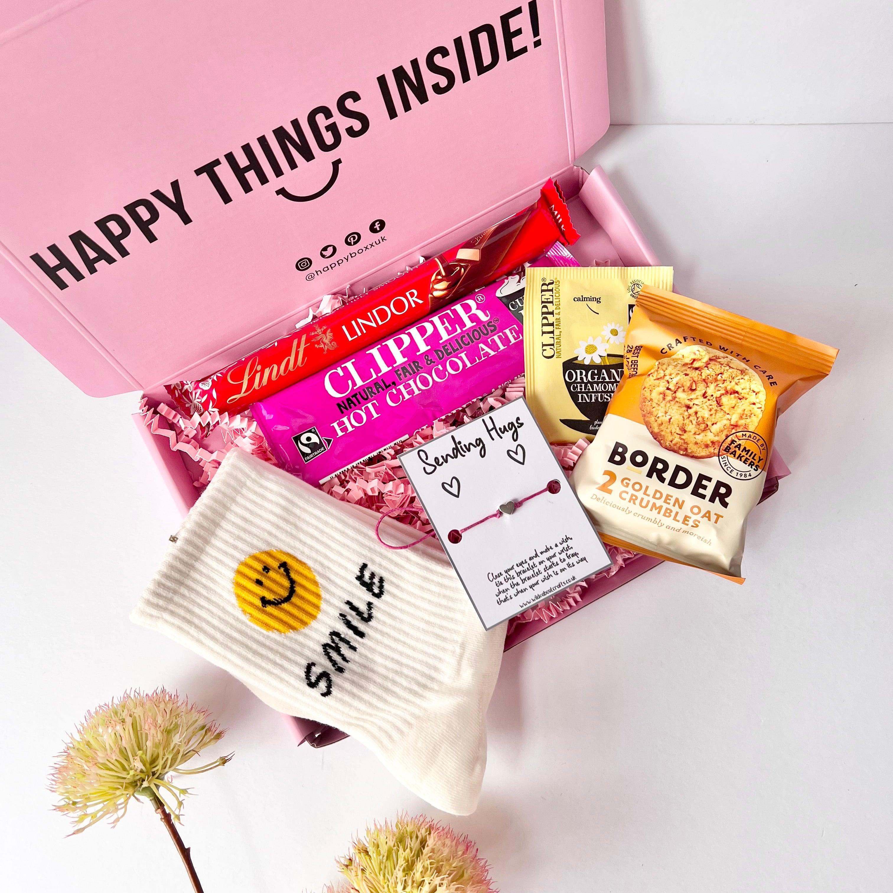 A pink gift box containing various items including a clippers hot chocolate sachet, a borders biscuit pack, a pair of smiley face crew socks, a sending hugs wish bracelet, a Lindor chocolate and a herbal tea bag.