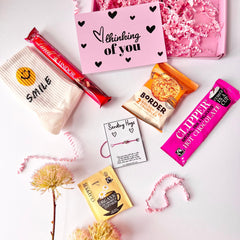 A flat lay image of clippers hot chocolate sachet, a borders biscuit pack, a pair of smiley face crew socks, a sending hugs wish bracelet, a Lindor chocolate, a herbal tea bag and a 'Thinking of you' greeting card.