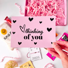 A pink greeting card with the text 'Thinking of you' and black illustrated love hearts print all over.