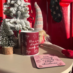 Red mug with Christmas text on a table with a small Christmas tree and decorations in the background.
