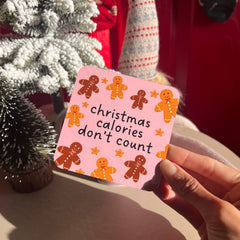 Hand holding a pink card with gingerbread men and 'Christmas calories don't count' text in front of a Christmas tree.