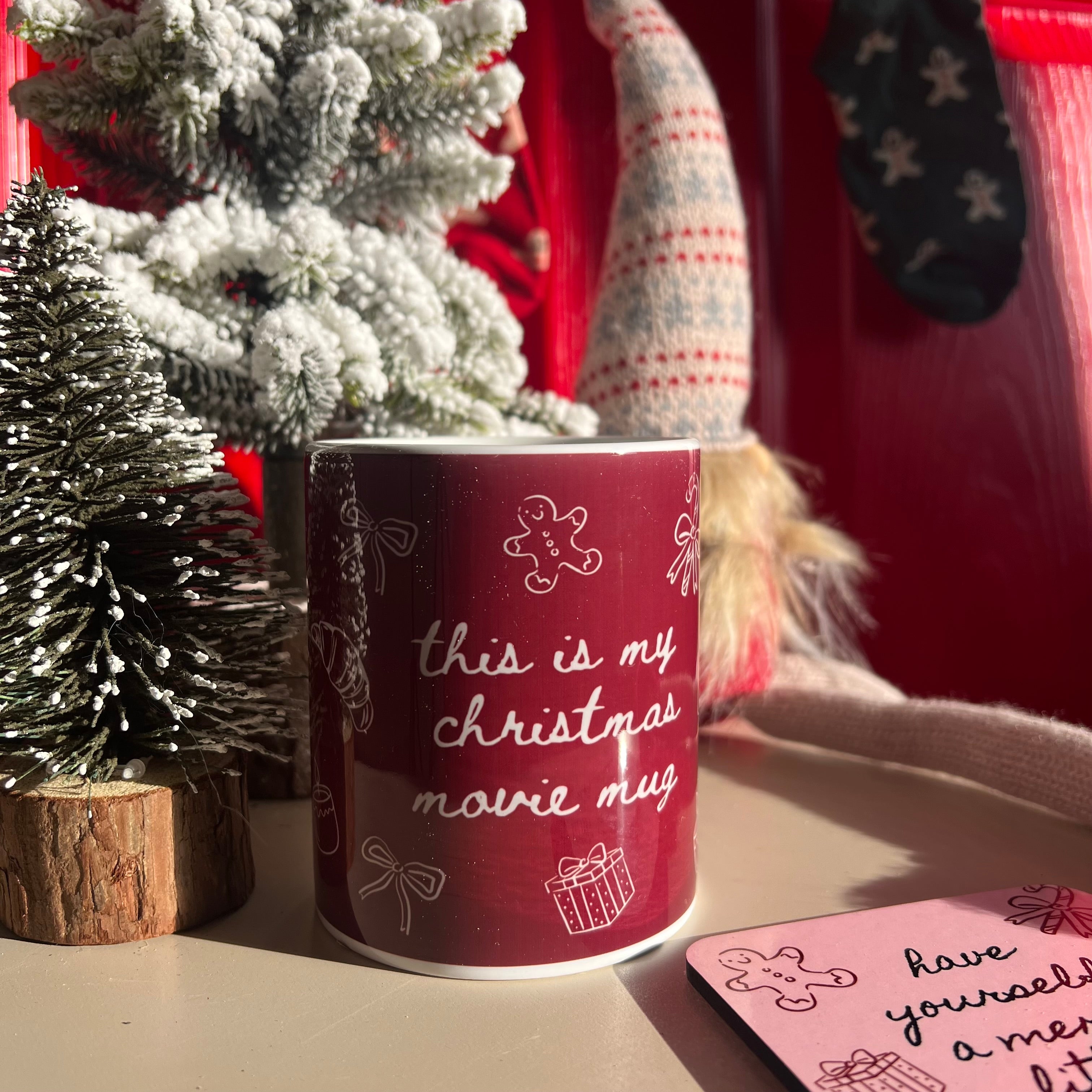 Red mug with Christmas-themed text in a festive setting with a tree and stockings.