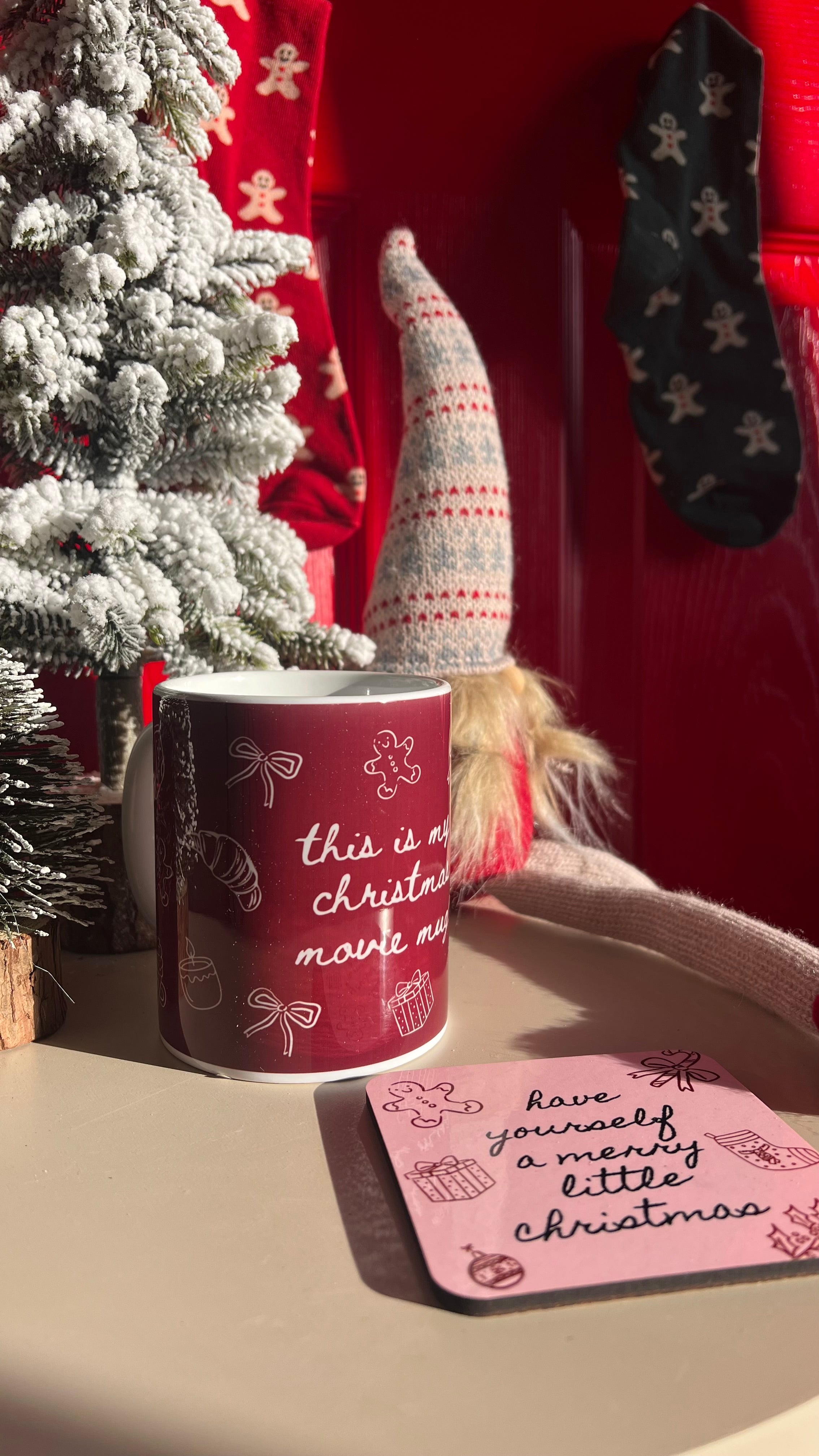 Red mug with Christmas text on a table next to a small tree and decorative items.