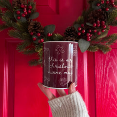 Red mug with 'this is my Christmas movie mug' text held in front of a red door with festive decorations.