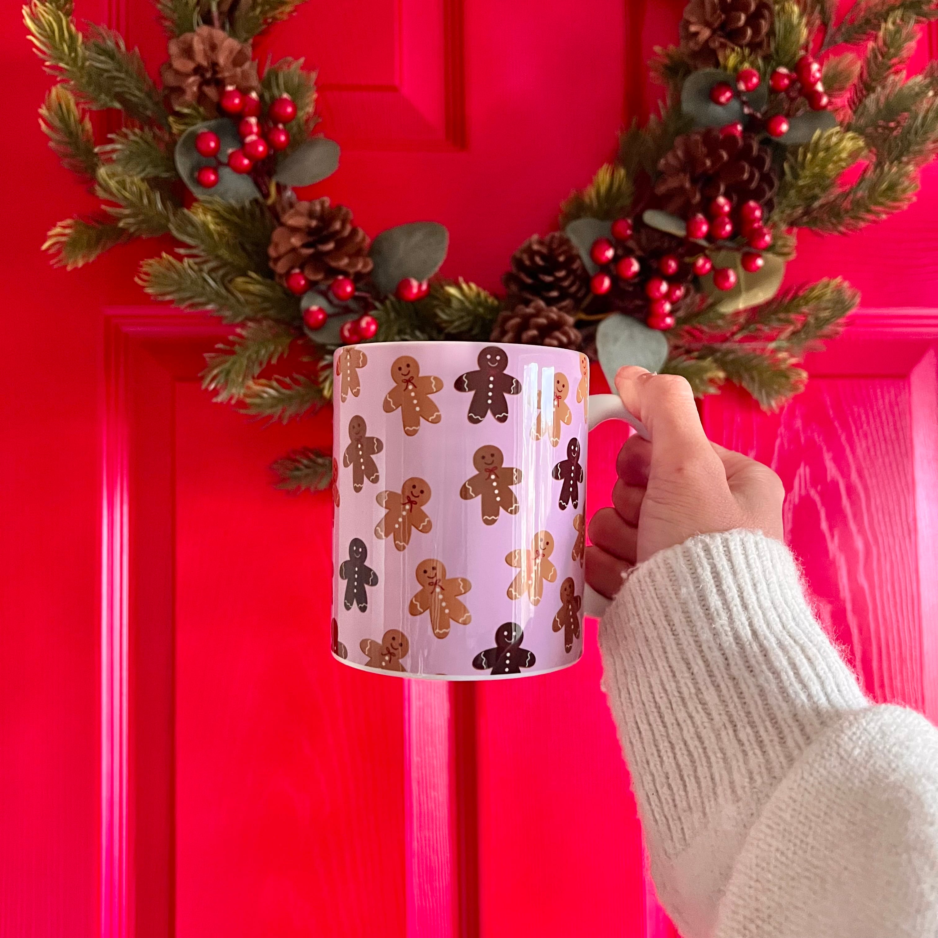 Hand holding a mug with gingerbread man pattern in front of a red door with a wreath.