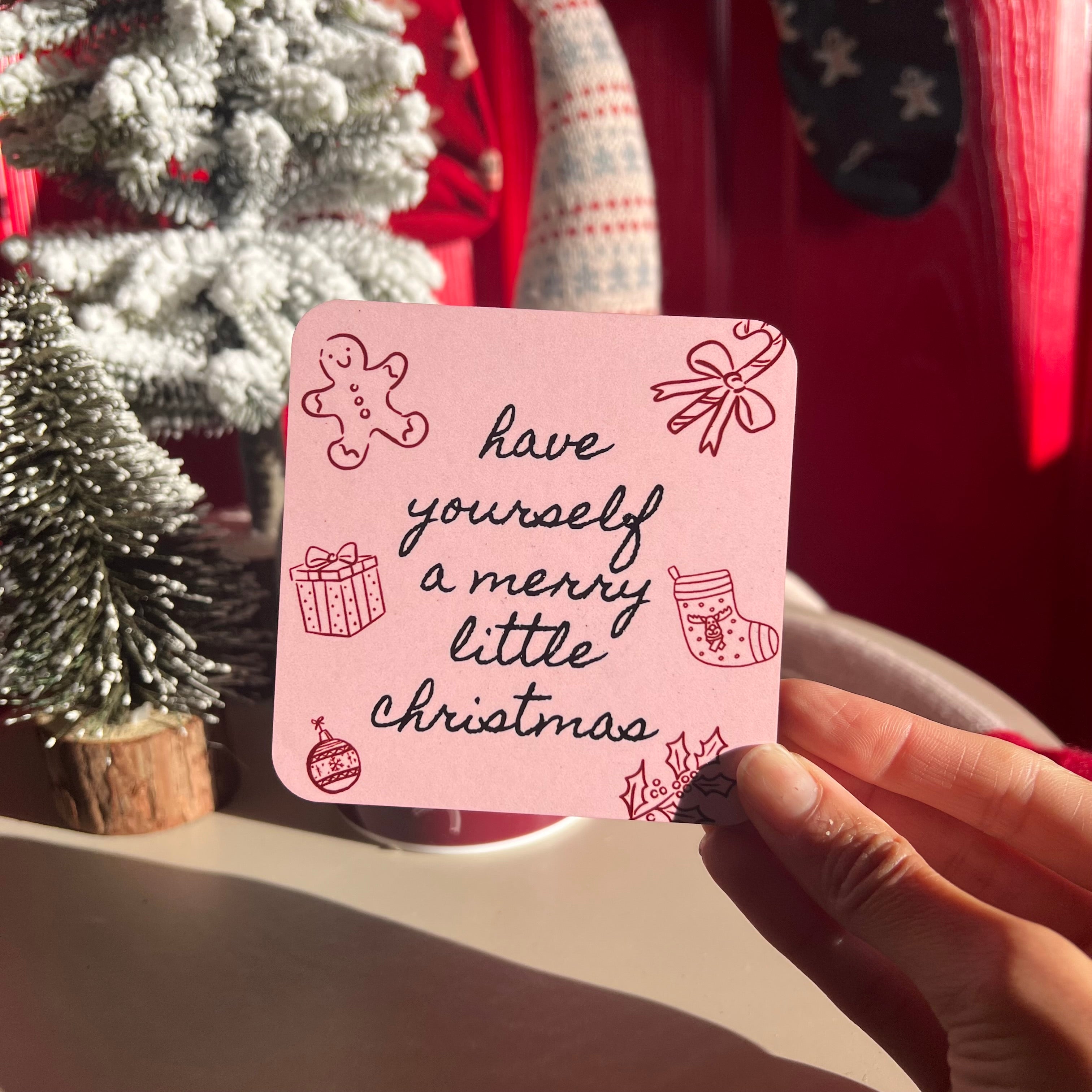 Hand holding a pink card with 'have yourself a merry little Christmas' text and festive illustrations against a Christmas-themed background.