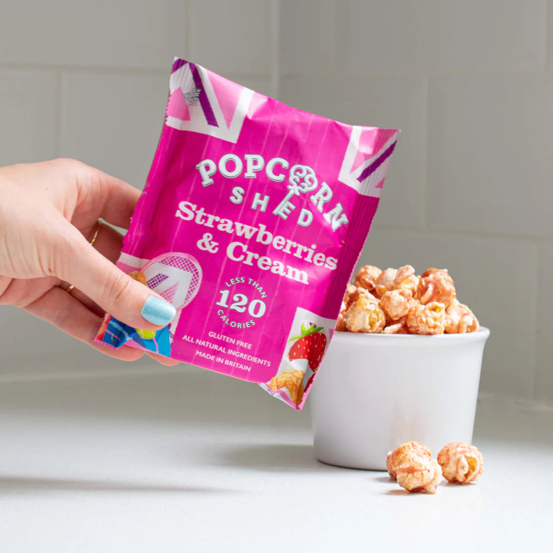 Hand holding a pink 'Popcorn Shed Strawberries & Cream' packet with popcorn in a bowl on a white surface.